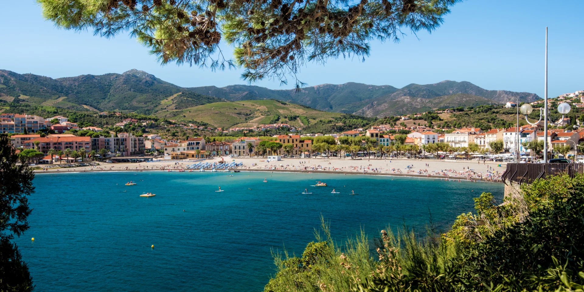 Vue panoramique sur la plage principale bordée d’immeubles et de collines verdoyantes, avec la mer turquoise au premier plan.
