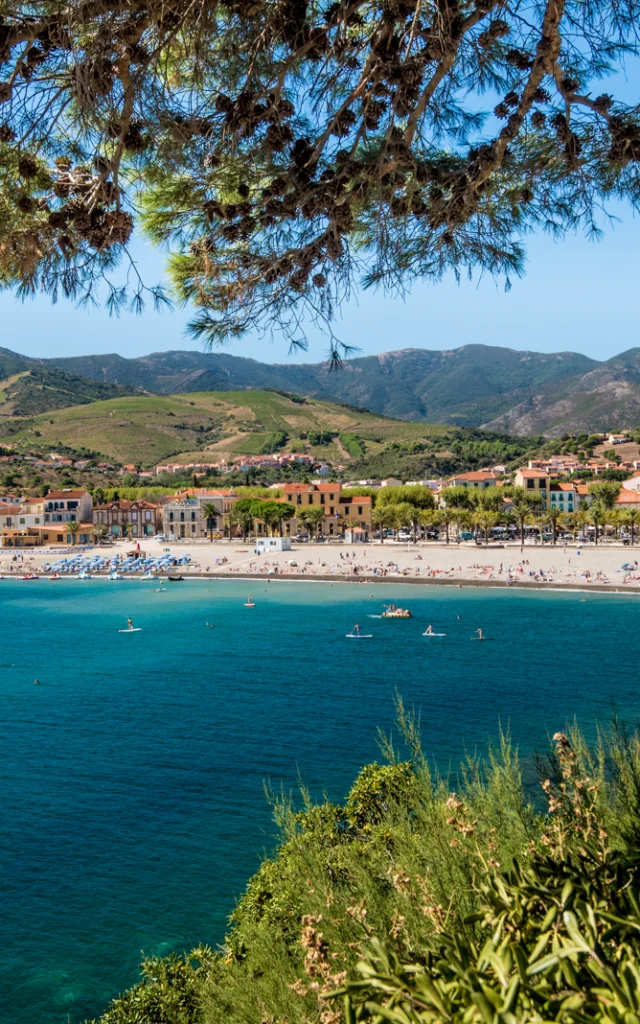 Vue panoramique sur la plage principale bordée d’immeubles et de collines verdoyantes, avec la mer turquoise au premier plan.