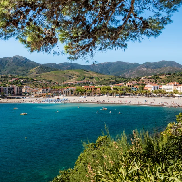 Vue panoramique sur la plage principale bordée d’immeubles et de collines verdoyantes, avec la mer turquoise au premier plan.