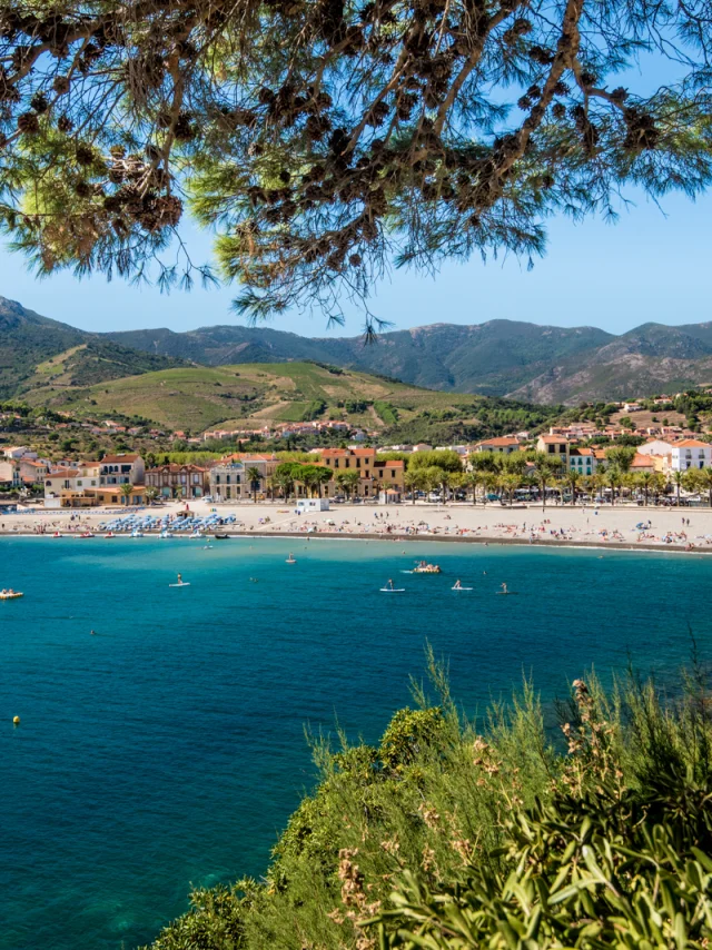 Vue panoramique sur la plage principale bordée d’immeubles et de collines verdoyantes, avec la mer turquoise au premier plan.
