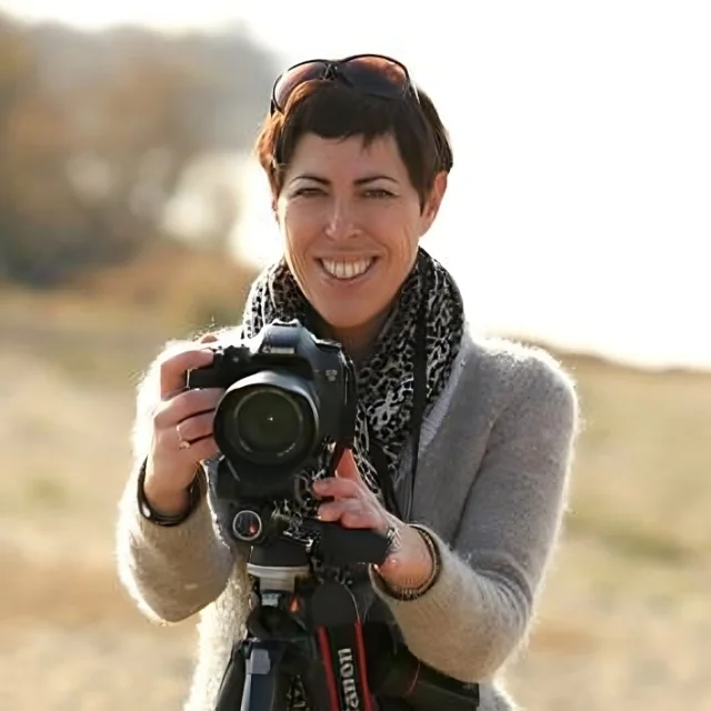 Portrait d’une femme souriante tenant un appareil photo sur trépied en extérieur, dans une lumière douce.