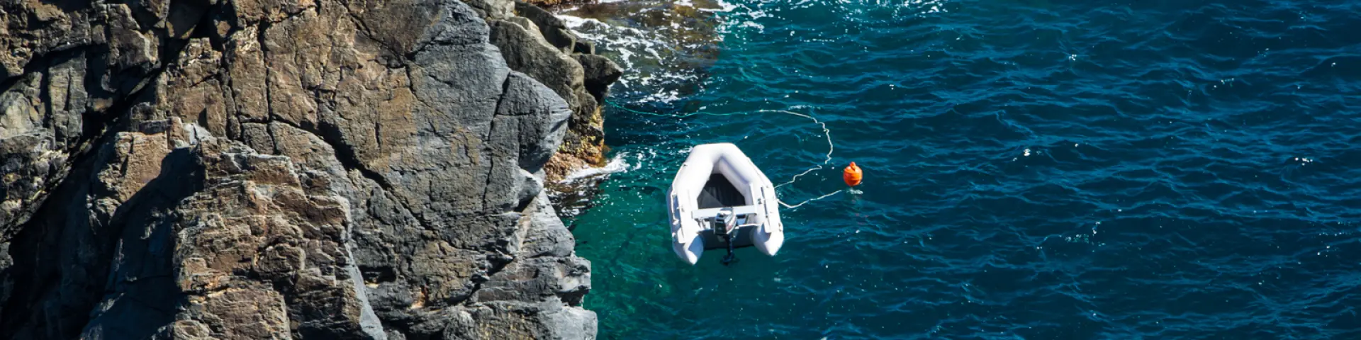Vue aérienne d’un petit bateau gonflable amarré près des rochers, sur une mer d’un bleu profond aux reflets turquoise.