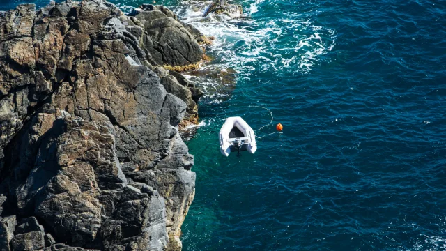 Vue aérienne d’un petit bateau gonflable amarré près des rochers, sur une mer d’un bleu profond aux reflets turquoise.