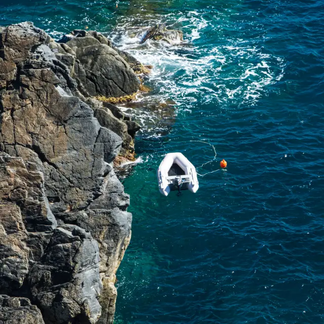 Vue aérienne d’un petit bateau gonflable amarré près des rochers, sur une mer d’un bleu profond aux reflets turquoise.