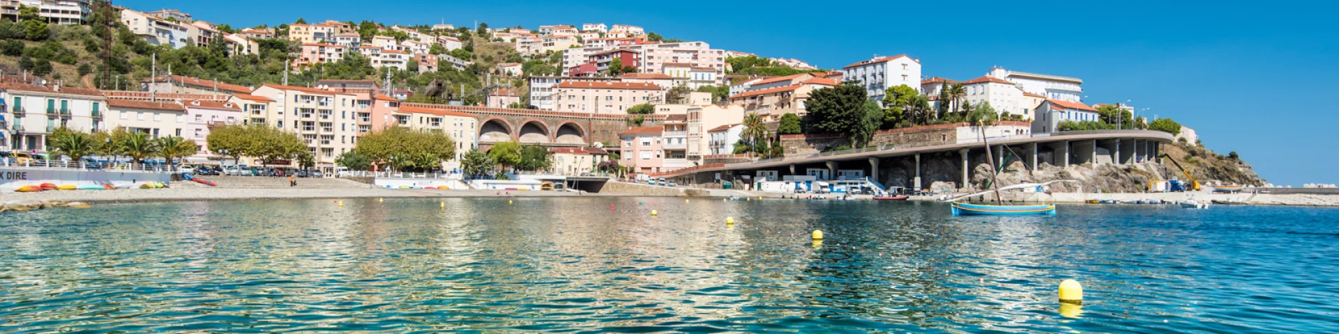 Vue depuis la mer sur la station balnéaire nichée à flanc de colline, avec ses maisons colorées, ses arcs de viaduc et l’eau turquoise du littoral méditerranéen.