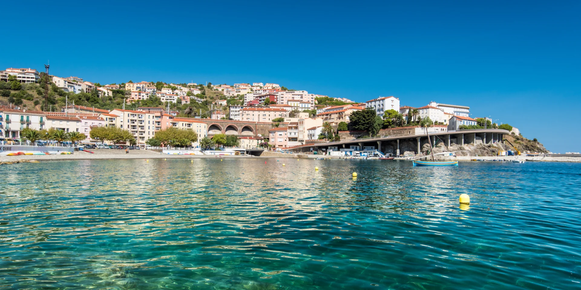 Vue depuis la mer sur la station balnéaire nichée à flanc de colline, avec ses maisons colorées, ses arcs de viaduc et l’eau turquoise du littoral méditerranéen.