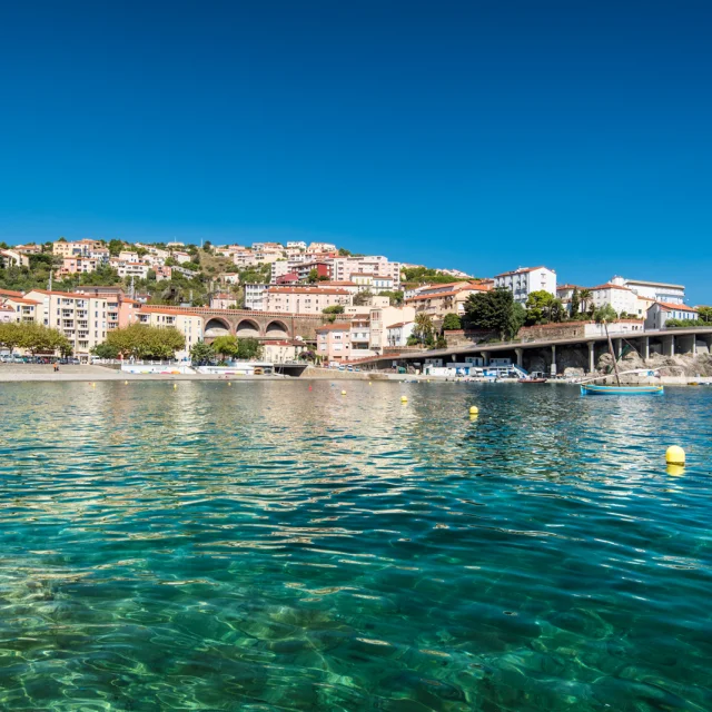 Vue depuis la mer sur la station balnéaire nichée à flanc de colline, avec ses maisons colorées, ses arcs de viaduc et l’eau turquoise du littoral méditerranéen.