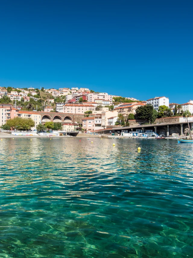 Vue depuis la mer sur la station balnéaire nichée à flanc de colline, avec ses maisons colorées, ses arcs de viaduc et l’eau turquoise du littoral méditerranéen.