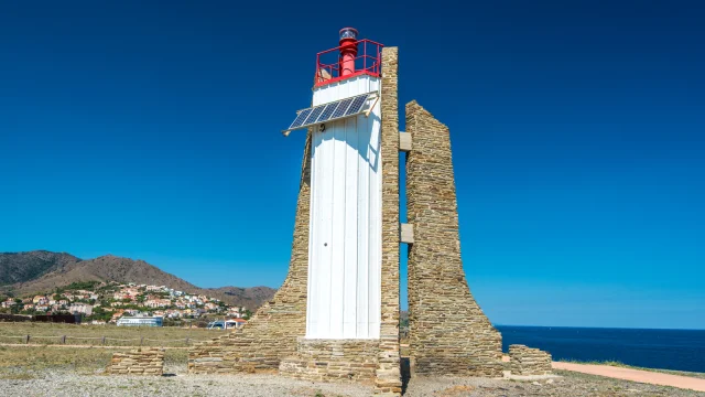 Phare moderne en pierre et métal surplombant la mer Méditerranée, avec les collines et le village en arrière-plan sous un ciel bleu.