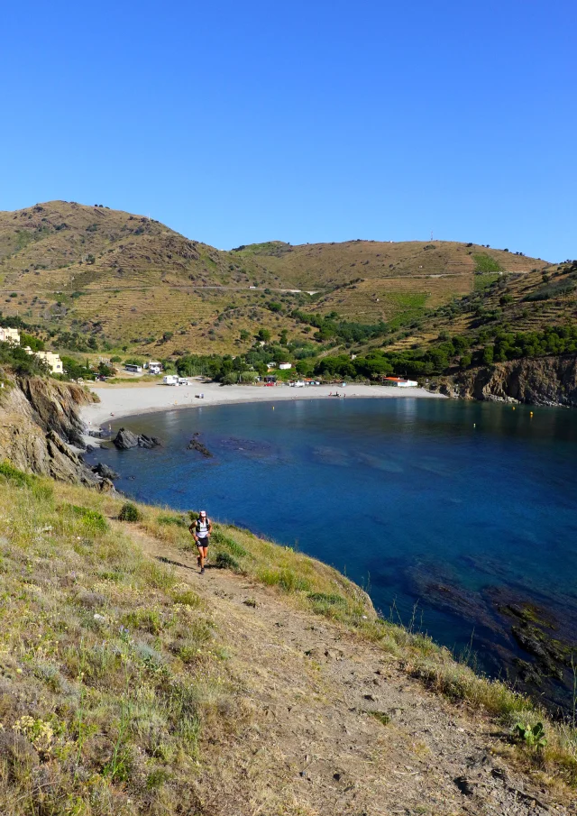 Randonneur sur un sentier dominant une crique aux eaux bleues, entourée de collines verdoyantes sur la Côte Vermeille.