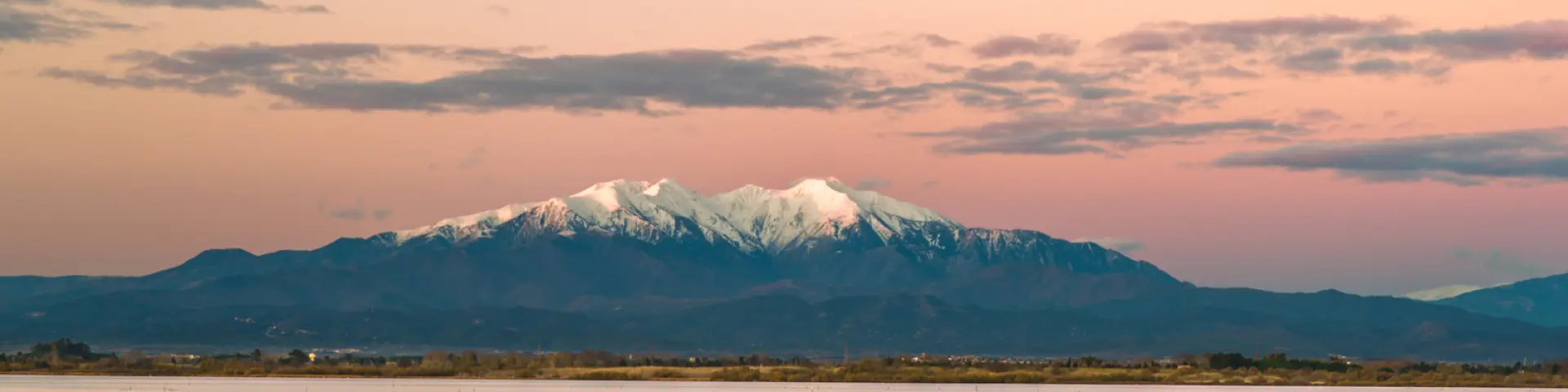 Petit bateau bleu flottant sur un étang au coucher du soleil, avec le Canigó enneigé en arrière-plan sous un ciel rosé.