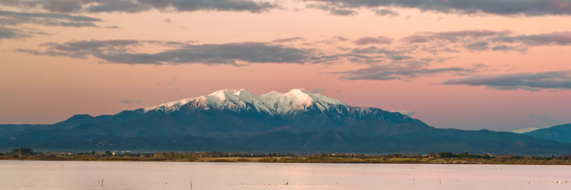 Petit bateau bleu flottant sur un étang au coucher du soleil, avec le Canigó enneigé en arrière-plan sous un ciel rosé.