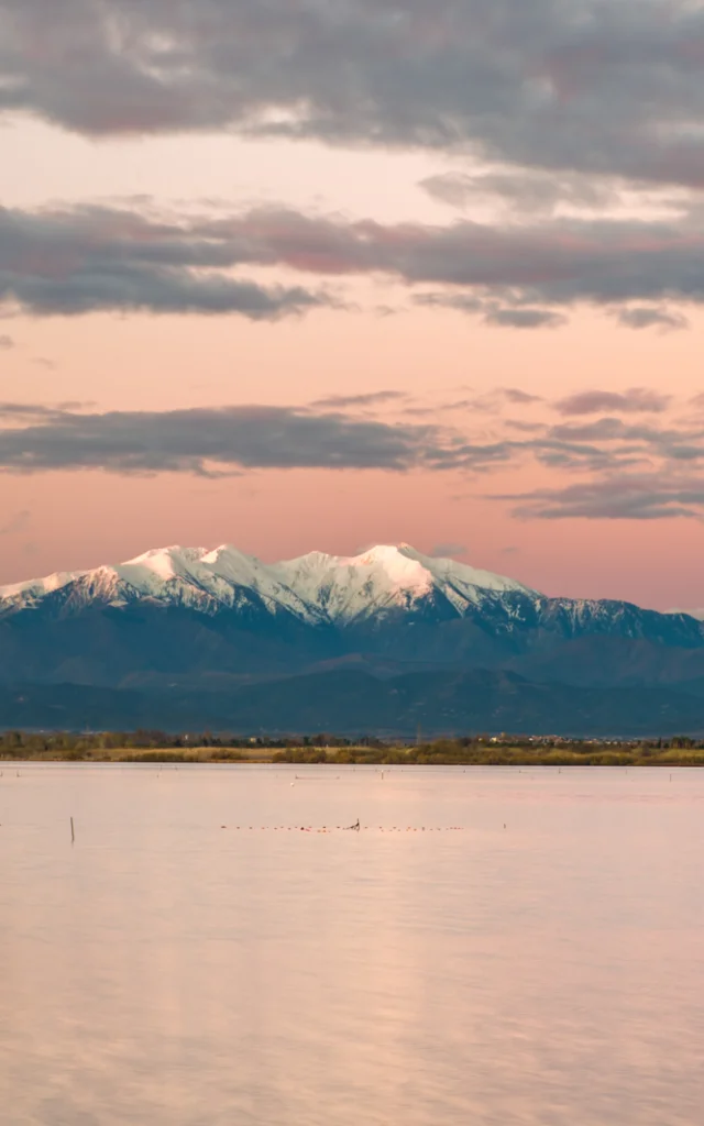 Petit bateau bleu flottant sur un étang au coucher du soleil, avec le Canigó enneigé en arrière-plan sous un ciel rosé.