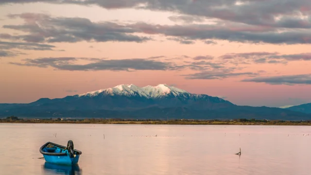 Petit bateau bleu flottant sur un étang au coucher du soleil, avec le Canigó enneigé en arrière-plan sous un ciel rosé.