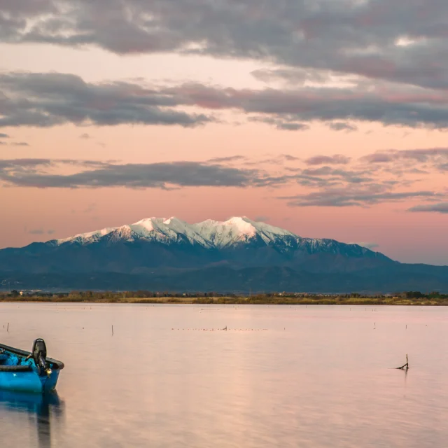 Petit bateau bleu flottant sur un étang au coucher du soleil, avec le Canigó enneigé en arrière-plan sous un ciel rosé.