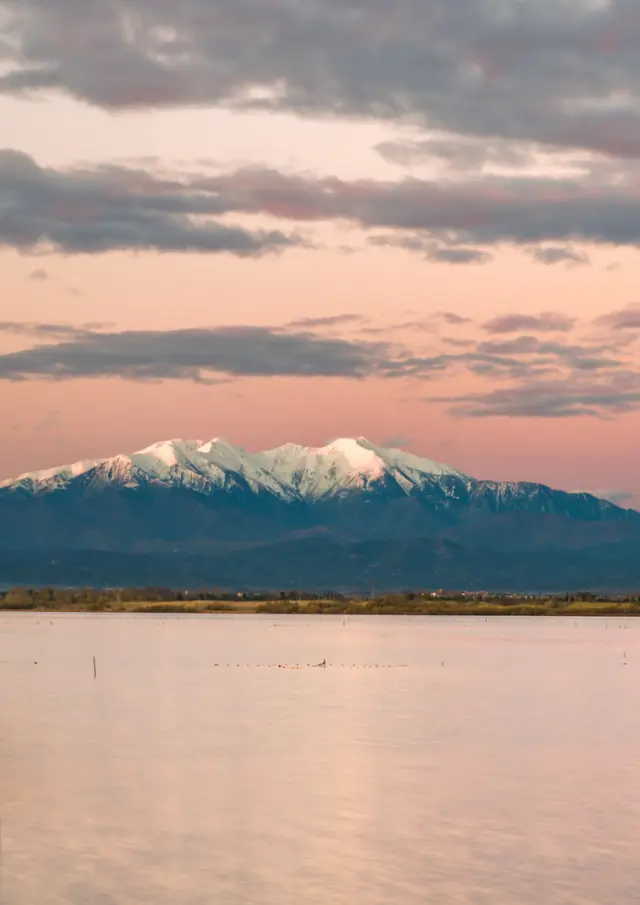 Petit bateau bleu flottant sur un étang au coucher du soleil, avec le Canigó enneigé en arrière-plan sous un ciel rosé.