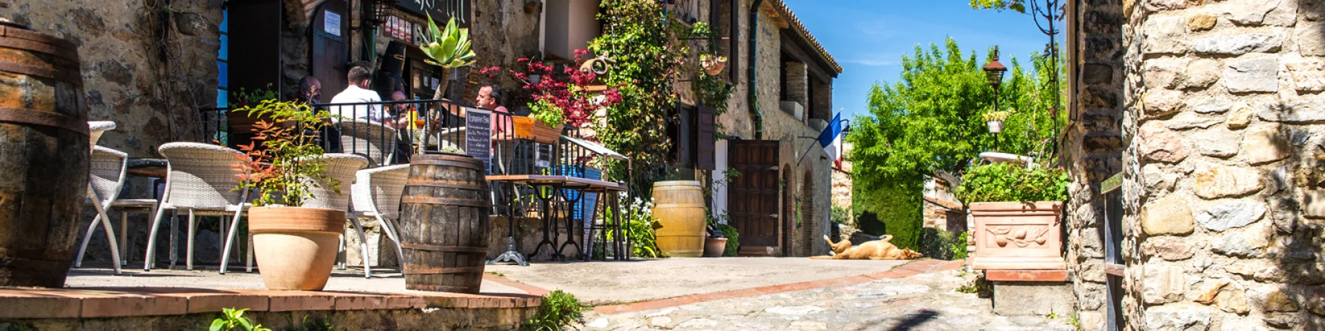 Ruelle pavée bordée de maisons en pierre avec une terrasse fleurie de restaurant, sous un ciel bleu ensoleillé.