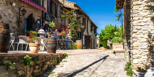 Ruelle pavée bordée de maisons en pierre avec une terrasse fleurie de restaurant, sous un ciel bleu ensoleillé.
