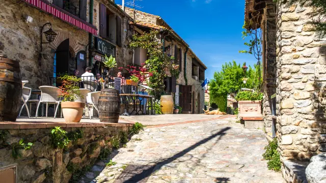 Ruelle pavée bordée de maisons en pierre avec une terrasse fleurie de restaurant, sous un ciel bleu ensoleillé.