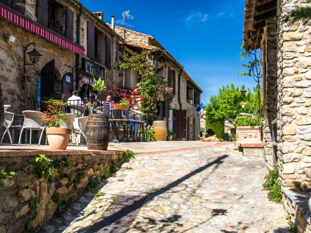 Ruelle pavée bordée de maisons en pierre avec une terrasse fleurie de restaurant, sous un ciel bleu ensoleillé.