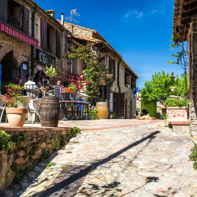 Ruelle pavée bordée de maisons en pierre avec une terrasse fleurie de restaurant, sous un ciel bleu ensoleillé.