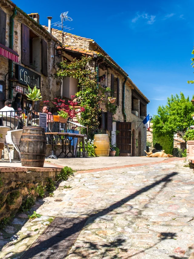 Ruelle pavée bordée de maisons en pierre avec une terrasse fleurie de restaurant, sous un ciel bleu ensoleillé.