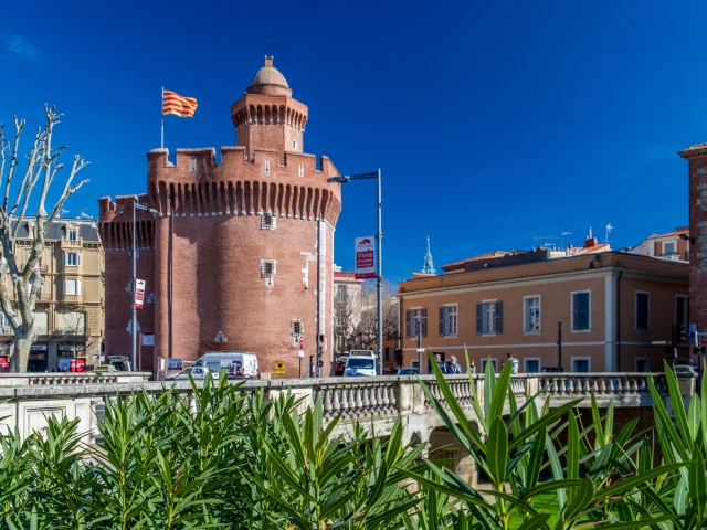 Le Castillet de Perpignan, monument en briques rouges surmonté du drapeau catalan, sous un ciel bleu.