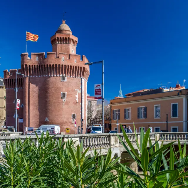 Le Castillet de Perpignan, monument en briques rouges surmonté du drapeau catalan, sous un ciel bleu.