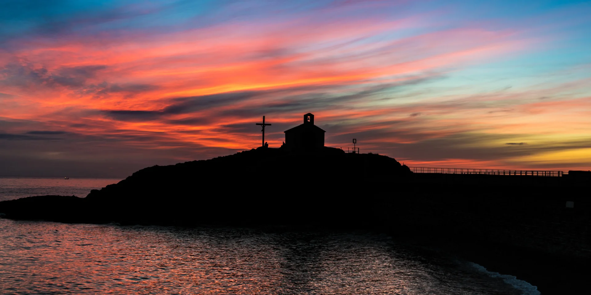 Silhouette d’une chapelle et d’une croix se détachant au coucher du soleil sur un ciel aux teintes orange, rose et bleue, en bord de mer.