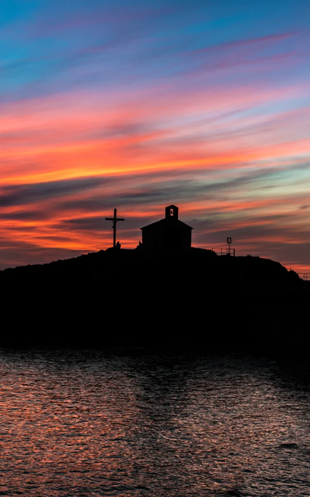 Silhouette d’une chapelle et d’une croix se détachant au coucher du soleil sur un ciel aux teintes orange, rose et bleue, en bord de mer.