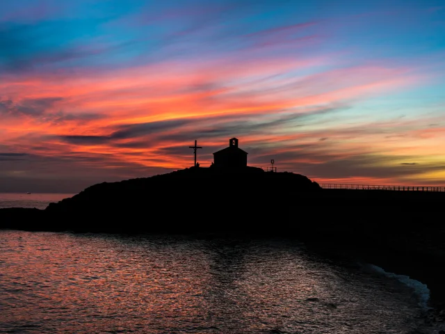 Silhouette d’une chapelle et d’une croix se détachant au coucher du soleil sur un ciel aux teintes orange, rose et bleue, en bord de mer.