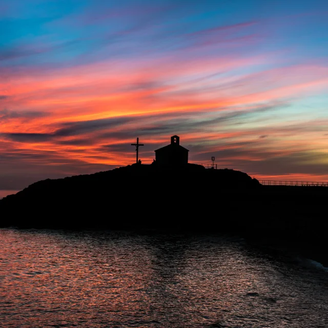 Silhouette d’une chapelle et d’une croix se détachant au coucher du soleil sur un ciel aux teintes orange, rose et bleue, en bord de mer.