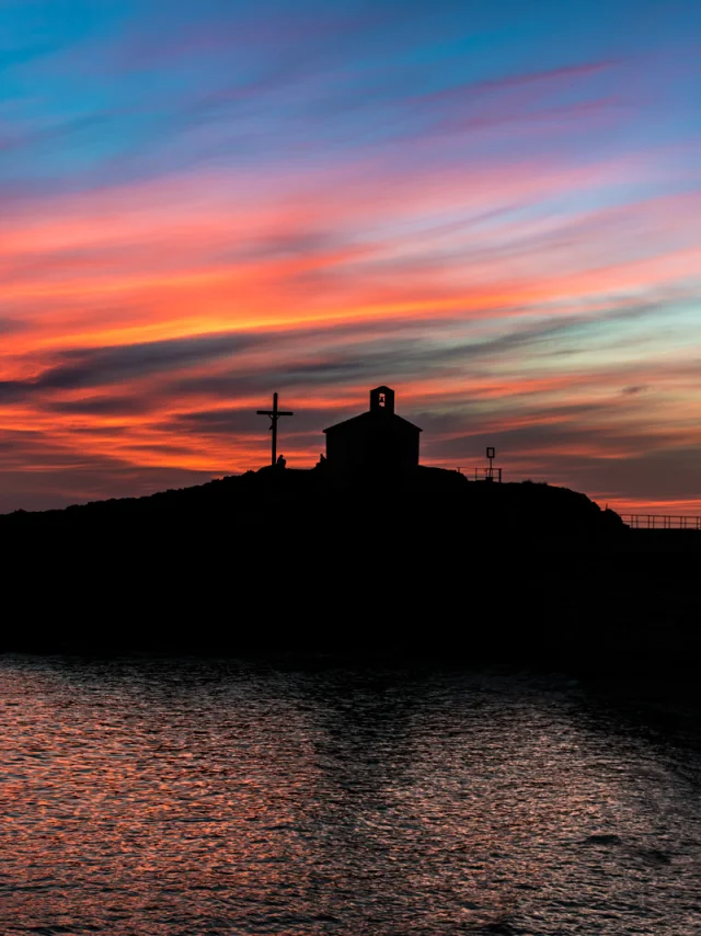 Silhouette d’une chapelle et d’une croix se détachant au coucher du soleil sur un ciel aux teintes orange, rose et bleue, en bord de mer.