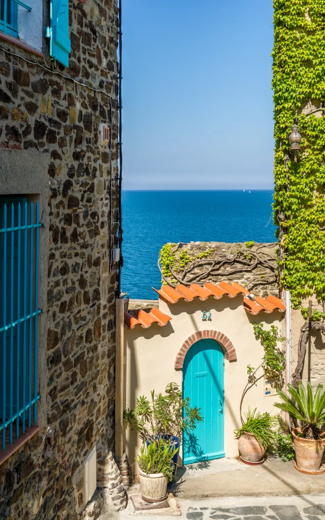 Petite ruelle en pierre menant à une maison aux volets et à la porte turquoise, avec vue sur la mer Méditerranée sous un ciel bleu.