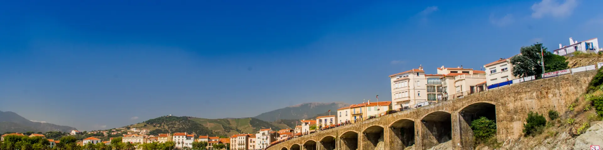 Deux personnes en kayak rouge pagaient au large d’une plage bordée d’immeubles et d’arcades en pierre, sous un ciel bleu éclatant.