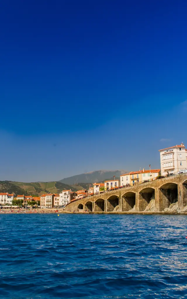 Deux personnes en kayak rouge pagaient au large d’une plage bordée d’immeubles et d’arcades en pierre, sous un ciel bleu éclatant.