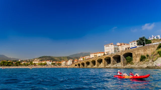 Deux personnes en kayak rouge pagaient au large d’une plage bordée d’immeubles et d’arcades en pierre, sous un ciel bleu éclatant.