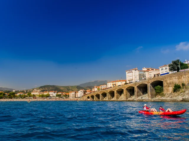 Deux personnes en kayak rouge pagaient au large d’une plage bordée d’immeubles et d’arcades en pierre, sous un ciel bleu éclatant.