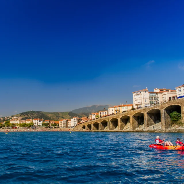 Deux personnes en kayak rouge pagaient au large d’une plage bordée d’immeubles et d’arcades en pierre, sous un ciel bleu éclatant.