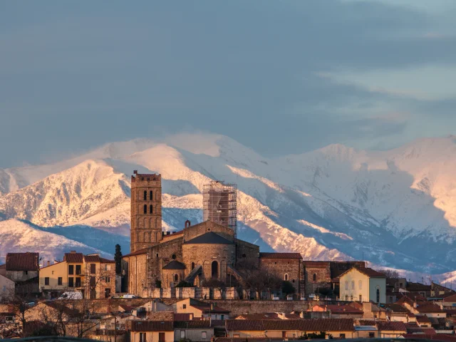 Vue sur la cathédrale d’Elne au coucher du soleil, avec en arrière-plan le massif du Canigó enneigé.