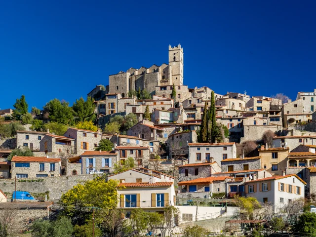 Vue du village d’Eus en Conflent, perché sur une colline et dominé par l’église Saint-Vincent, sous un ciel bleu intense des Pyrénées-Orientales.