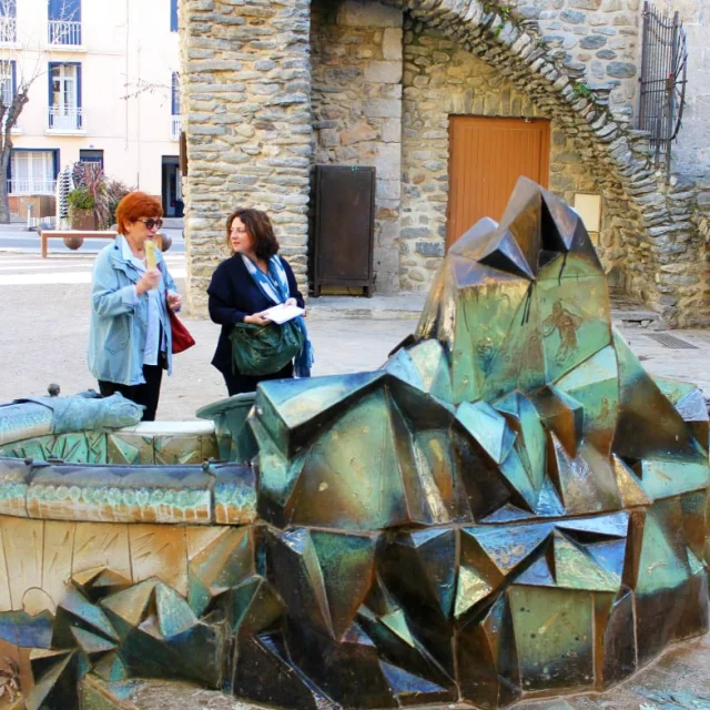 Deux femmes discutant devant la fontaine Dali à Céret, une œuvre en céramique aux formes géométriques et colorées, installée sur une place du centre historique.
