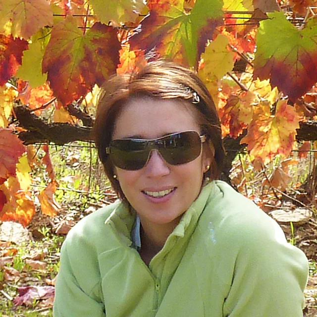 Personne souriante portant des lunettes de soleil et une veste verte, assise devant des vignes aux feuilles rouges et dorées en automne.