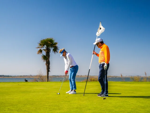 Deux golfeurs sur un green en bord de mer, sous un ciel bleu, l’un prêt à putter tandis que l’autre tient le drapeau du trou.