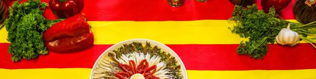 Assiette d’anchois de Collioure entourée de poivrons rouges et de persil, présentée sur une nappe aux couleurs catalanes rouge et jaune.