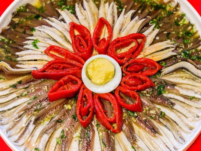 Assiette d’anchois de Collioure dressée en forme de fleur avec des lamelles de poivrons rouges et une touche d’aïoli au centre.