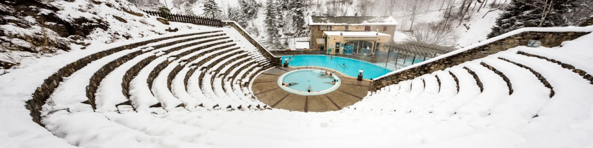 Vue en hauteur des bains de Saint-Thomas-les-Bains en hiver, avec des baigneurs profitant des bassins d’eau chaude naturelle entourés de neige.