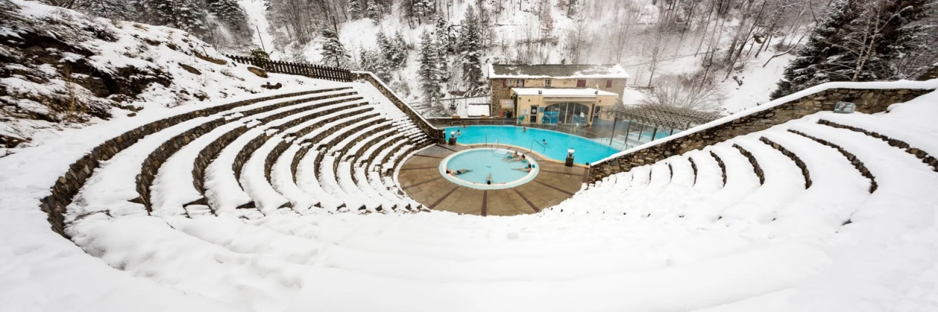 Vue en hauteur des bains de Saint-Thomas-les-Bains en hiver, avec des baigneurs profitant des bassins d’eau chaude naturelle entourés de neige.