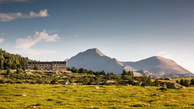 Grand bâtiment de montagne en pierre au pied des sommets du massif du Carlit, entouré d’un vaste plateau verdoyant dans les Pyrénées-Orientales.
