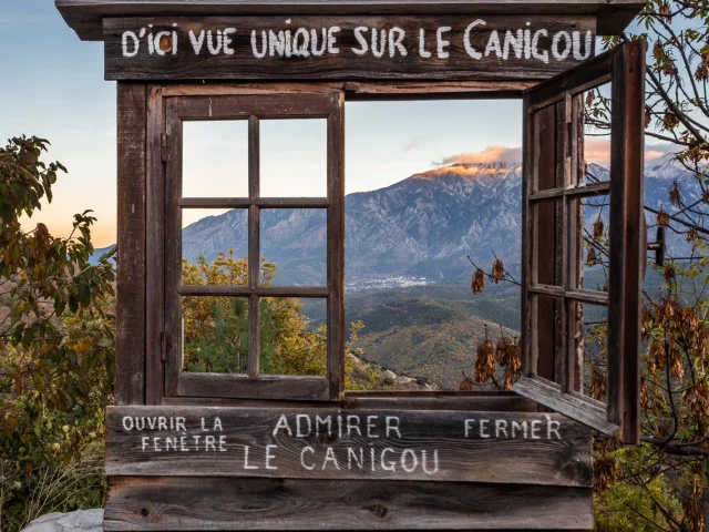 Cadre en bois en forme de fenêtre avec l’inscription « D’ici vue unique sur le Canigou », ouvrant sur une vue panoramique du mont Canigó au coucher du soleil.